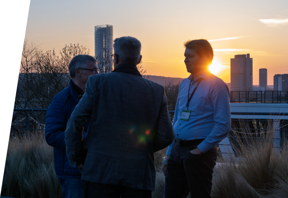 3 Personen, die auf der Dachterrasse stehen und sich unterhalten, mit Sonnenuntergang im Hintergrund.